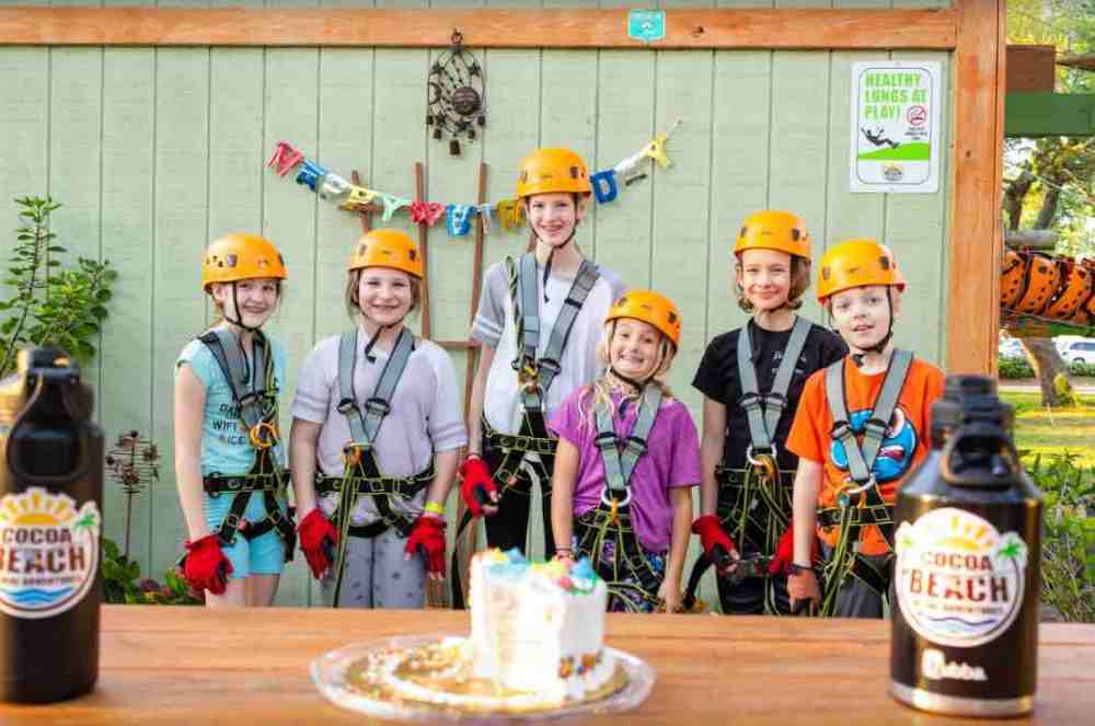Seven kids in helmets and gear pose for a group photo with a celebration banner and cake.