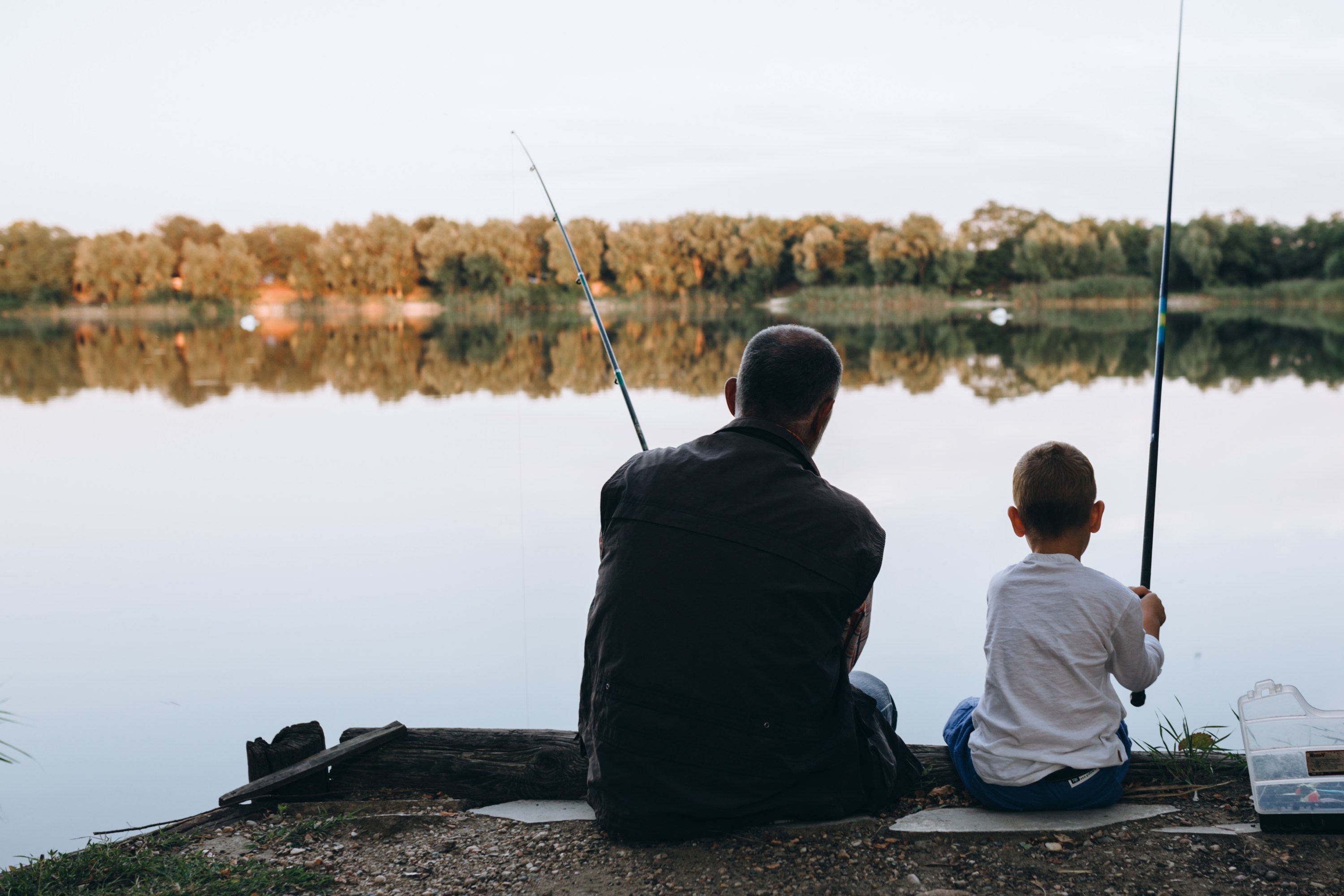 A man and child fishing by a calm lake with trees in the background.
