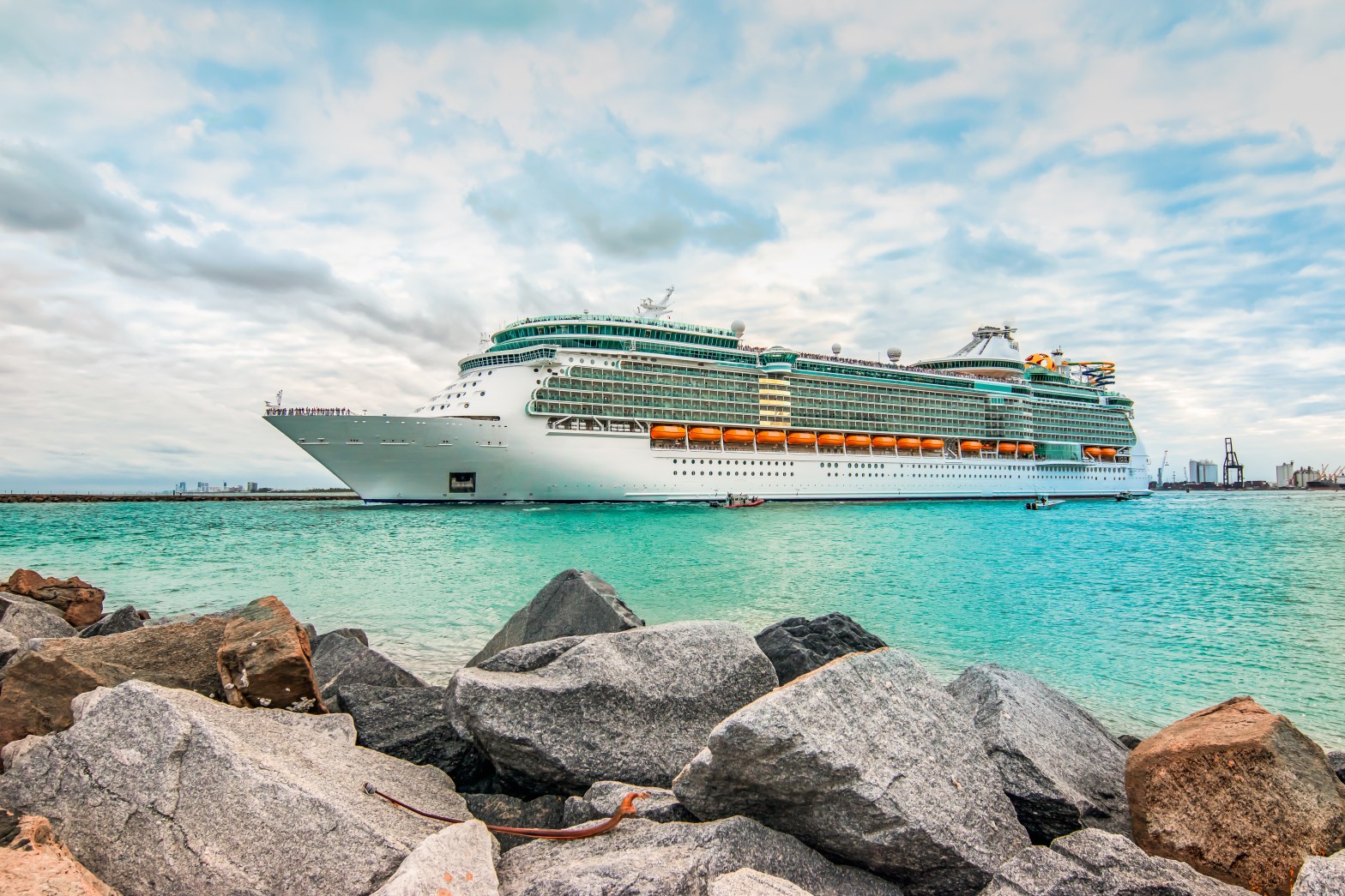 Large cruise ship on turquoise water with cloudy sky and rocks in foreground.