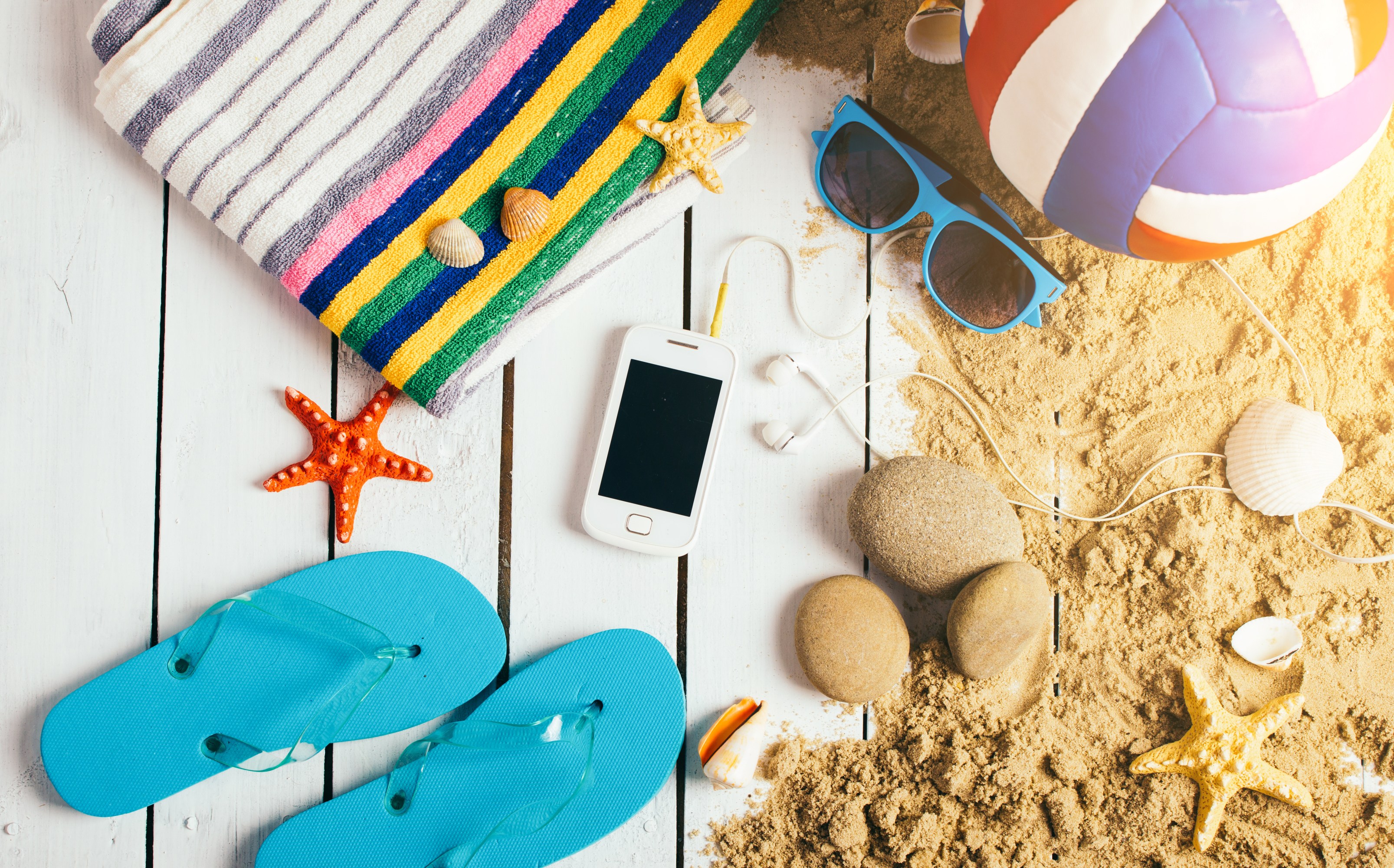 Beach essentials: striped towel, flip-flops, sunglasses, smartphone, volleyball, seashells, and sand on wood.