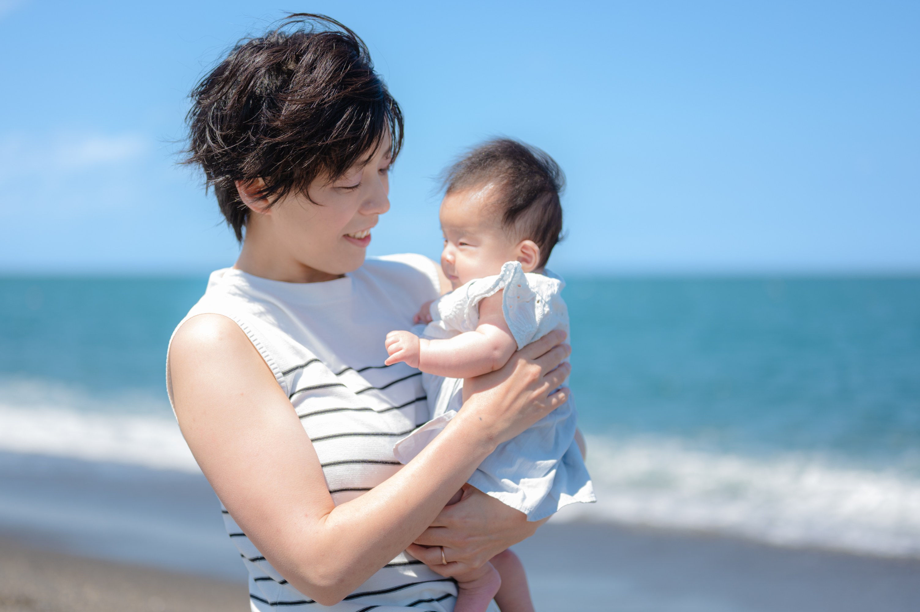 Woman holding a baby by the beach under a clear blue sky.