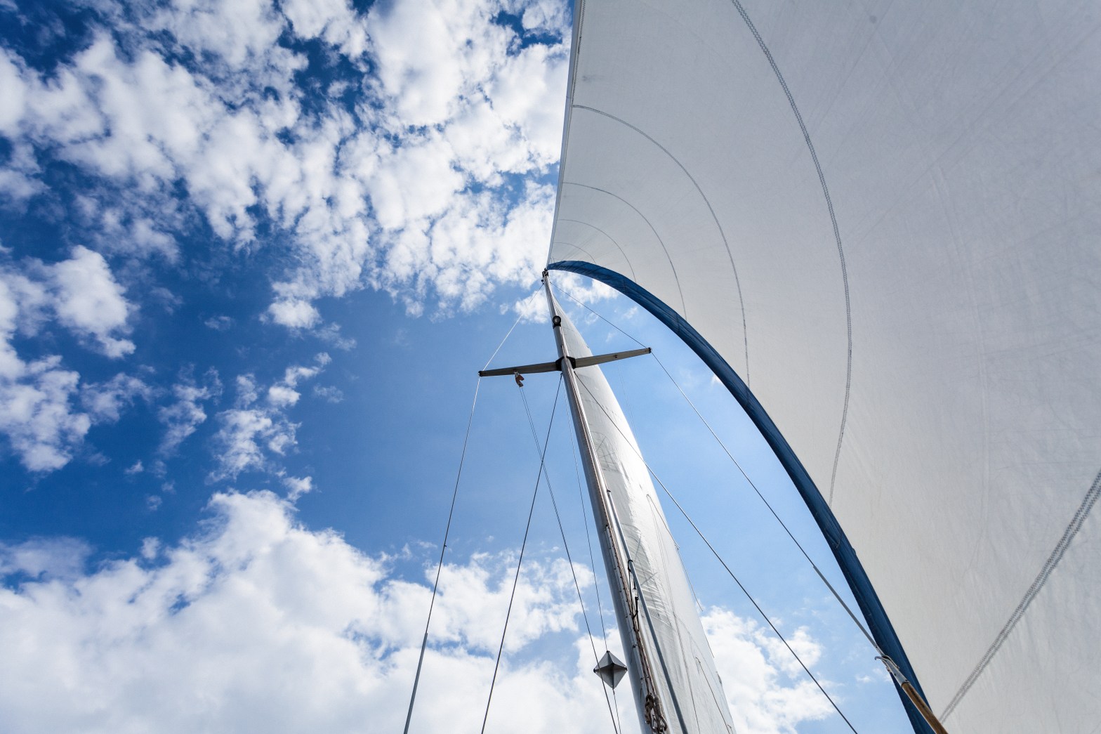 View up a sailboat mast with billowing white sails against a partly cloudy blue sky.