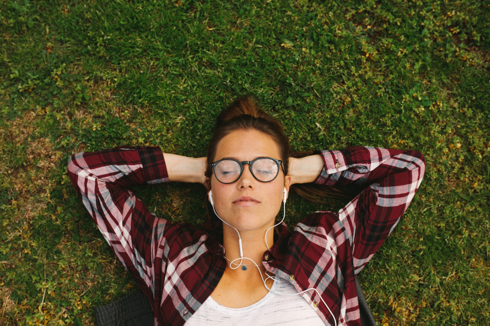 Person lying on grass, wearing glasses and headphones, eyes closed.