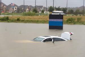 Car submerged in floodwater near a flooded area with houses and a sign partially visible.