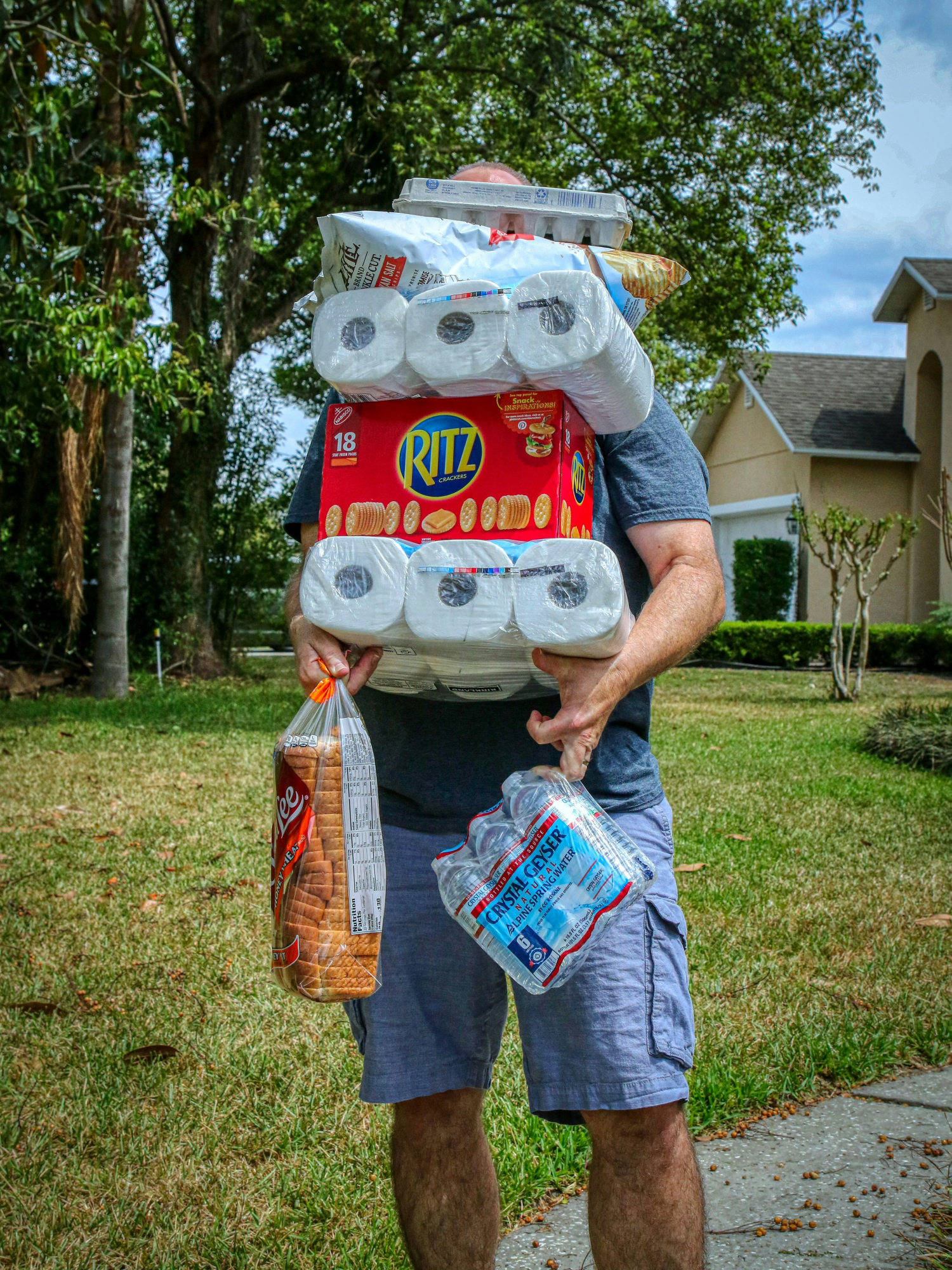 Person carries multiple grocery items, including toilet paper and snacks, outdoors.
