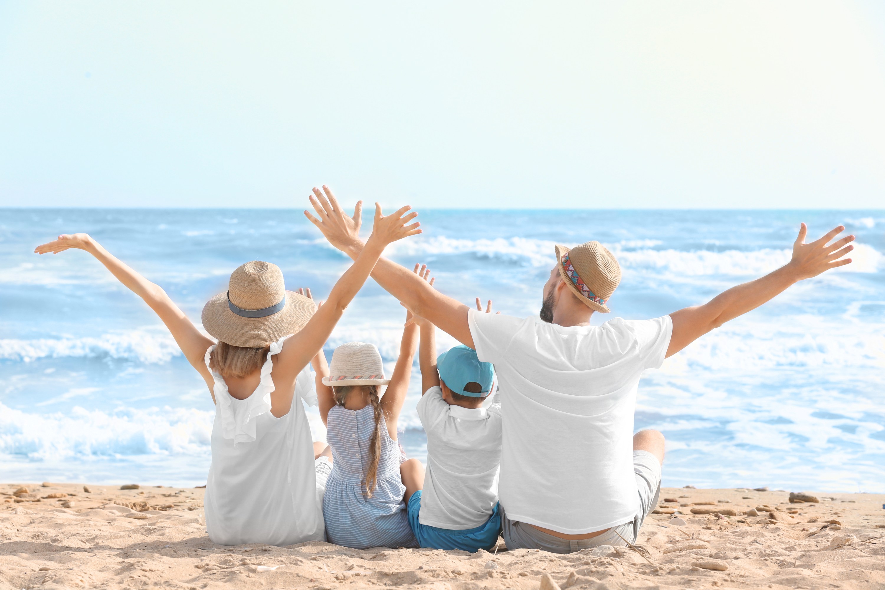 Family of four sitting on beach with arms raised, facing the ocean.