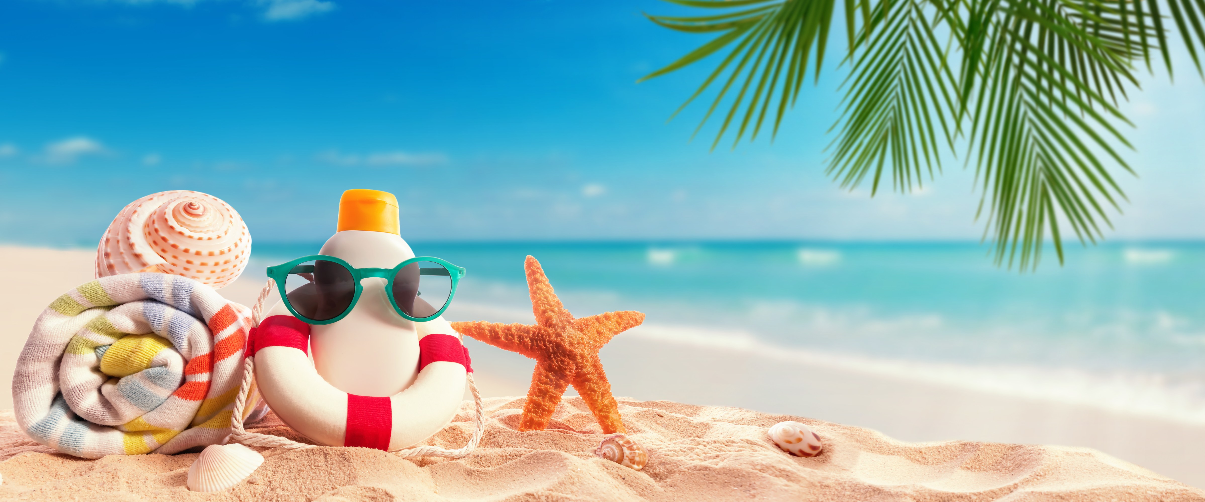 Beach scene with sunscreen, sunglasses, starfish, shells, and towel on sand, ocean and palm fronds in background.
