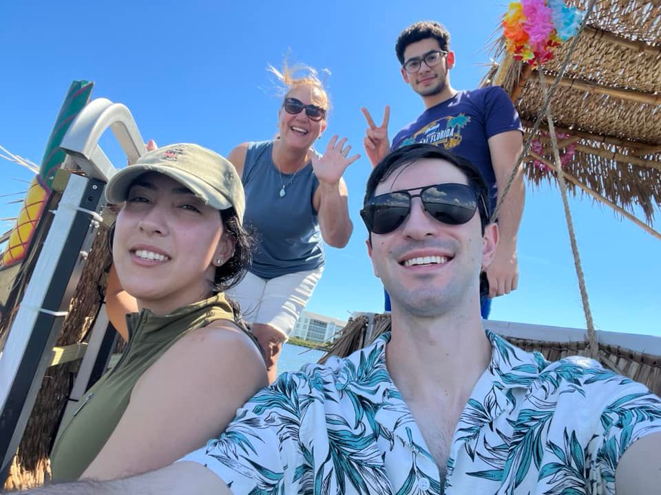 Four people smiling on a sunny day near a thatched structure by the water.