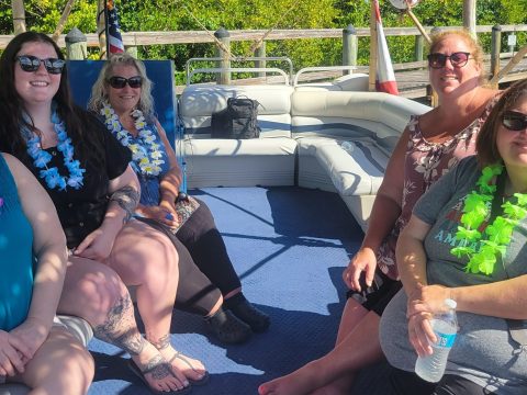 Five women wearing leis sitting on a boat under a thatched canopy, surrounded by greenery and water.