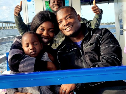 A smiling family on a boat, with one child asleep, blue sky background.