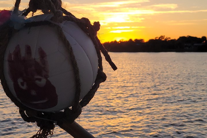 A decorated volleyball with a drawn face against a sunset over water.