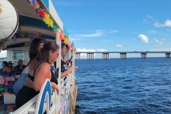 People on a boat enjoy a sunny day on the water, with a bridge in the background.