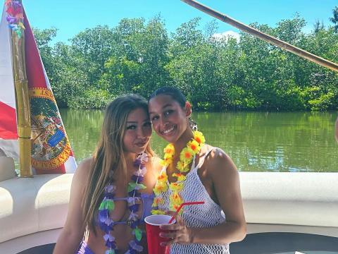 Two women with leis sitting on a boat, holding a red cup, with a tiki-style roof and green trees in the background.