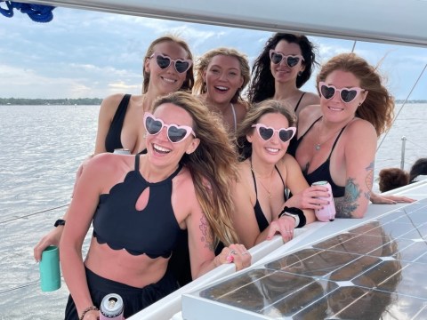 Six women in black swimwear with heart-shaped sunglasses smiling on a boat.