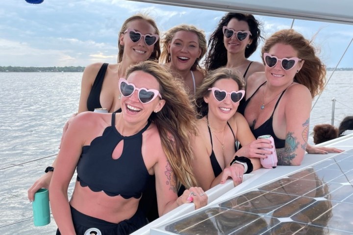 Six women in black swimwear with heart-shaped sunglasses smiling on a boat.