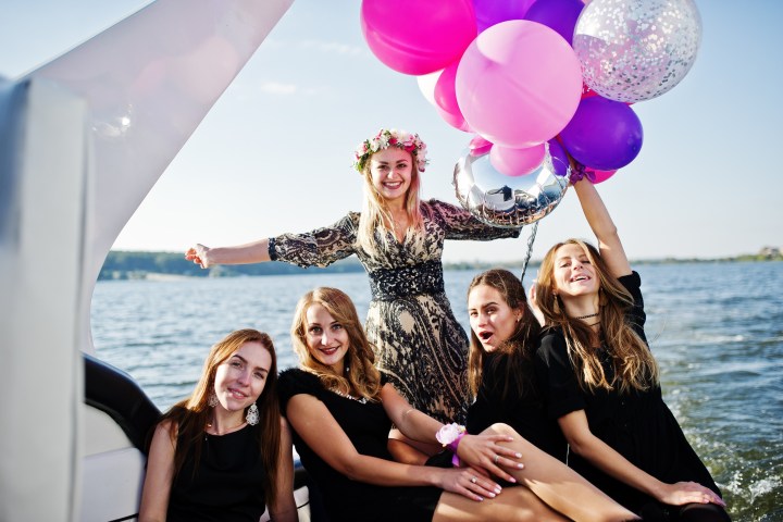 Five women on a boat with balloons, celebrating on a sunny day over water.