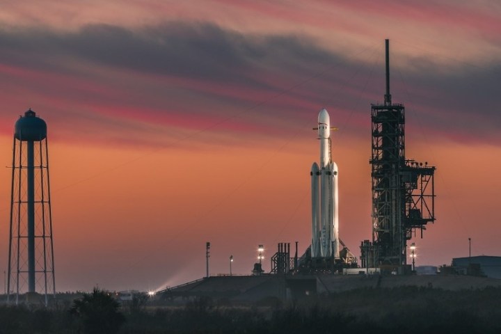 Rocket on launch pad at sunset next to a water tower, with orange sky and clouds.