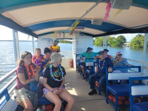 People wearing leis relax on a covered boat in a scenic waterway.