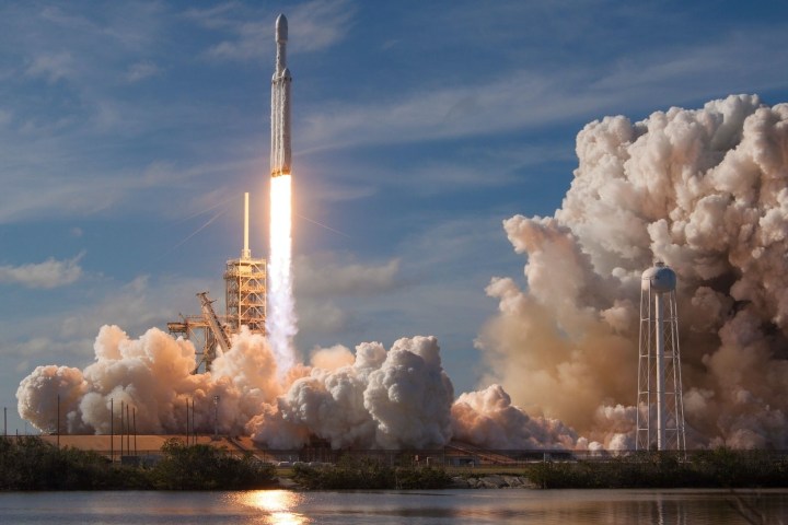 Rocket launching with flames and smoke, clear sky, water in foreground.