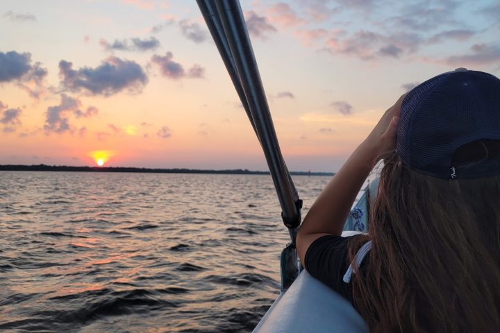 Person on boat photographing sunset over water with a hat on and vibrant sky.