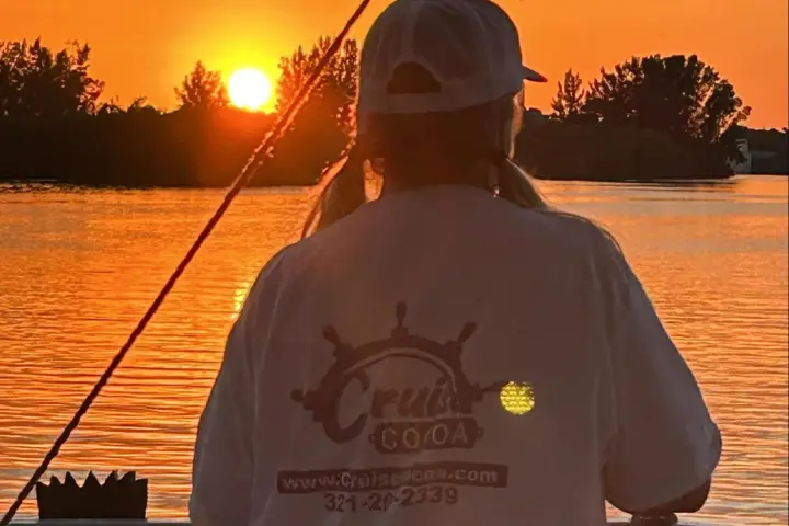 Person in cap watching sunset by a lake, wearing a shirt with a logo on the back.