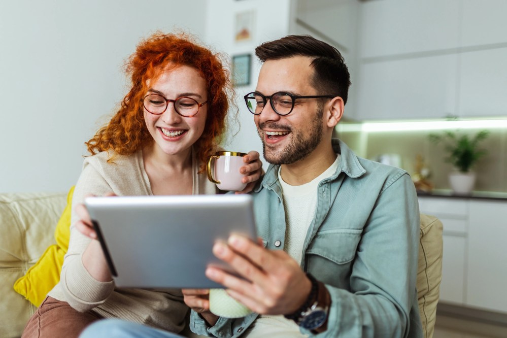Man and woman smiling at a tablet on a sofa, woman holding a mug.