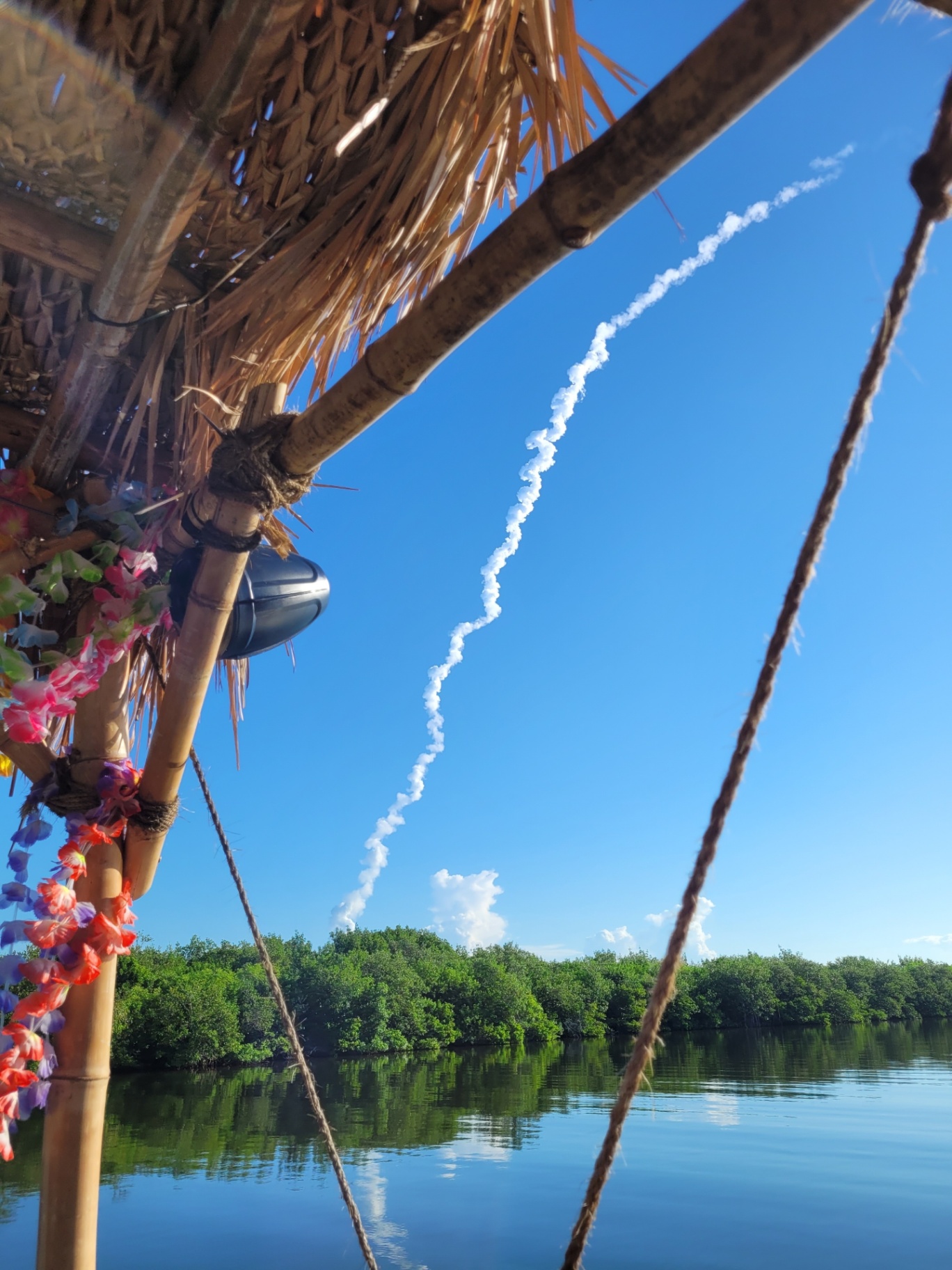 Rocket launch trail viewed from a thatched boat on a river with lush greenery.